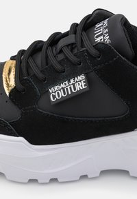 Baskets Versace Jeans Couture noires et dorées avec semelle blanche, lacets noirs, panneaux en daim et cuir, et logo de la marque visible sur le côté et la languette.