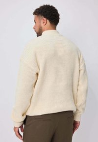Pull oversize couleur crème en tissu tricoté texturé, avec épaules tombantes et coupe décontractée, porté avec un pantalon vert olive.