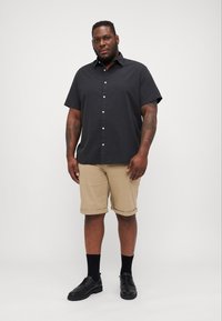 Chemise noire à manches courtes et à boutons, shorts beiges roulés, chaussures de ville noires et chaussettes noires. Tissu lisse, coupe décontractée. Design simple.