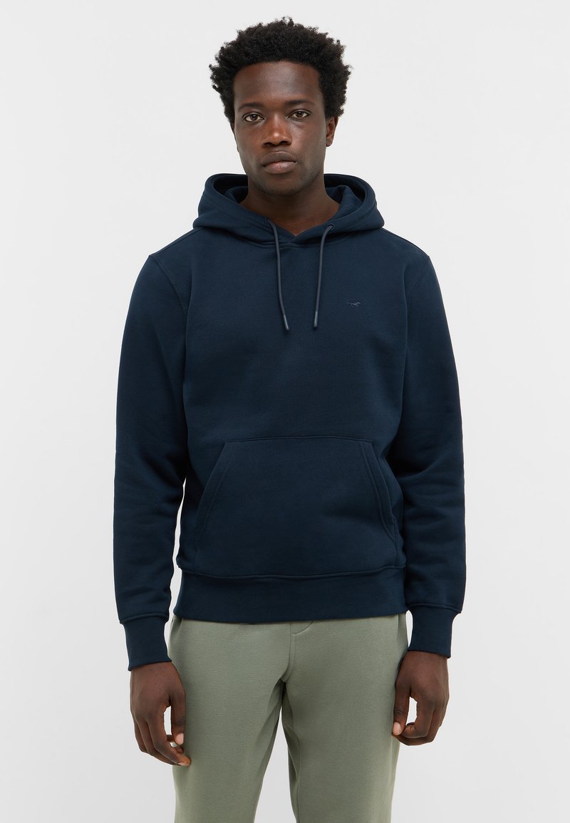 Młody czarnoskóry mężczyzna w granatowej bluzie z kapturem i jasnych zielonych spodniach, stojący z wyprostowanymi rękami na tle gładkiej białej ściany.