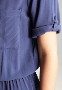 Marinblus med uppvikta ärmar, med en bröstficka. Mjuk, slät tyg med avslappnad passform och diskret lyster. Liten knappdetalj.