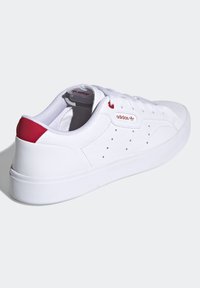 Vita sneakers med en slät läderöverdel, röd hälaccent och perforeringar för ventilation. Har vita snören och en gummisula.