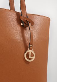 Braune strukturierte Kunstleder-Tasche mit einem goldenen Metall-Logo-Anhänger und schwarzem Akzentriemen, ausgestattet mit stabilen Griffen.