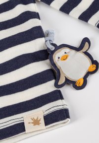 Navy und weiß gestrefter Stoff mit einem beigen Etikett mit einem Farbspritz-Logo. Befestigt ist ein plüschiger Pinguin-Schlüsselanhänger mit orangefarbenen Akzenten.