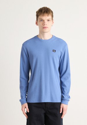 Light blue long-sleeve t-shirt in ribbed texture with round neckline, featuring a small navy logo patch on the left side.