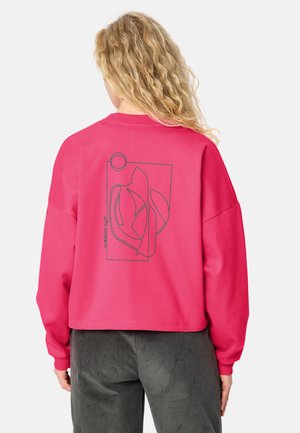 Person mit langen lockigen blonden Haaren trägt einen leuchtend pinken Pullover mit abstrakter Linienkunst und dem Text "outdoor 24/7" auf dem Rücken, kombiniert mit dunklen Hosen.