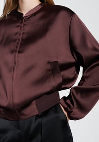 Giacca bomber di raso bordeaux con colletto tondo, chiusura a zip e tasca laterale. Presenta maniche arricciate e orlo a coste.