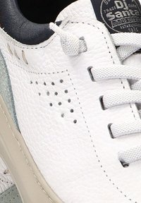 Zapatilla blanca de cuero con un acabado texturizado, acento de ante gris, cordones blancos planos y detalles perforados en el lateral. Suela de color claro.