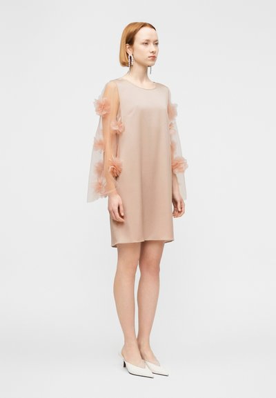 Φόρεμα ανοιχτό ροζ με διάφανα μανίκια διακοσμημένα με υφασμάτινα λουλούδια. Απλός σχεδιασμός σε γραμμή Α, λεία υφή και μήκος έως το γόνατο. Φορεμένο με λευκά παπούτσια.