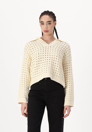 Cream, knitted sweater with a v-neck, loose fit, and a pattern of small holes. Long sleeves with ribbed cuffs, paired with black pants.