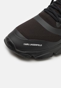 Svart sportskor med meshnät, gummisula och texturerade detaljer. Har en logotyptryck på sidan och en strömlinjeformad design.