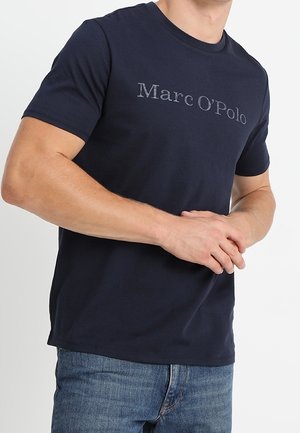 Man draagt een marineblauw Marc O'Polo T-shirt en blauwe spijkerbroek, handen samengevouwen bij de taille, tegen een effen witte achtergrond.