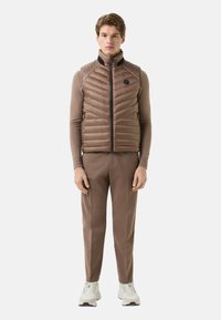 Braune Steppweste mit Chevron-Steppung, hohem Kragen, Frontreißverschluss und Seitentaschen. Kombiniert mit passenden braunen, maßgeschneiderten Hosen und weißen Sneakern.