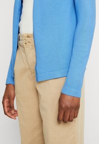 Ljusblå cardigan med texturerad stickning, med rak skärning och ribbade manschetter, parat med beige byxor och markerade fingrar.