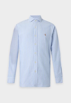 Giovane uomo che indossa una camicia a righe azzurro chiaro con bottoni, jeans chiari a gamba larga, sneakers bianche, in piedi con una mano in tasca.