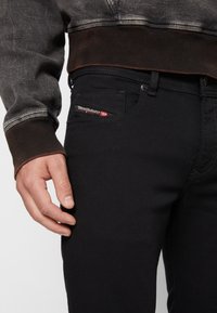Jeans de mezclilla negra con un diseño ajustado. Presentan una etiqueta de logo roja en el bolsillo frontal y un suéter gris texturizado visible arriba.