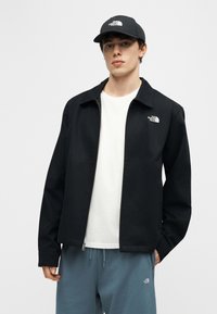 Homme portant une veste zippée noire, un T-shirt blanc, un pantalon bleu à cordon et une casquette noire, tous avec le logo The North Face.