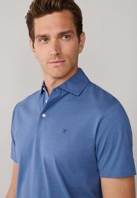 Polo à manches courtes bleu moyen, fabriqué en tissu lisse. Doté d'un col boutonné et d'un petit emblème brodé sur la poitrine gauche.