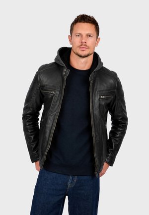 Homme portant une veste en cuir noire avec une capuche grise, un pull bleu marine et un jean bleu, debout les mains dans les poches devant un fond uni.