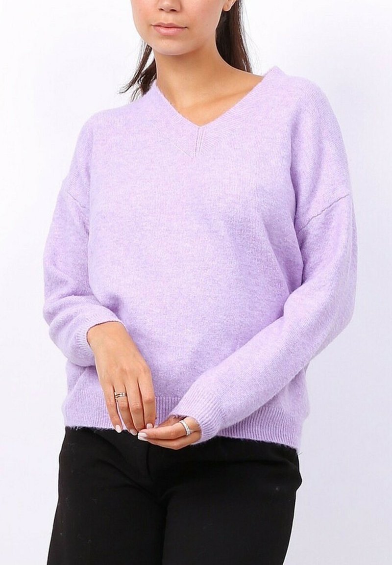 Pull en lilas à col en V, coupe ample, manches longues et poignets côtelés. Texture douce et design décontracté. Porté par-dessus un pantalon noir.