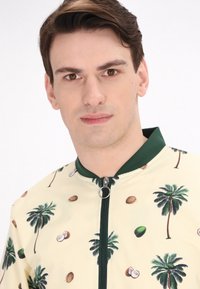 Lichtgele jas met palmbomen en kokosnootprint, groene ribbels op de kraag en een frontzipper met een ronde trekhaak. Zachte textuur.