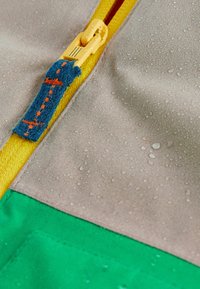 La chaqueta resistente al agua presenta un diseño multicolor con una parte superior gris, una cremallera amarilla y una sección inferior verde. Se pueden ver gotas de agua sobre el material.