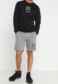 Chemise à manches longues noire avec logo blanc, associée à un short gris avec logo. Tissu texturé. Chaussures de sport noires avec lacets.