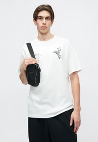 Biela tričko s grafickým potlačou a čierne šortky, drží malú čiernu crossbody tašku so zipsom a logom.