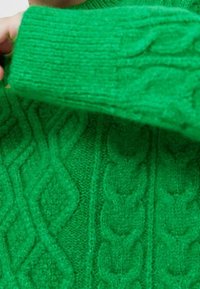 Pull vert en maille avec motifs torsadés, tissu texturé et poignet côtelé. Gros plan sur les détails de la manche.