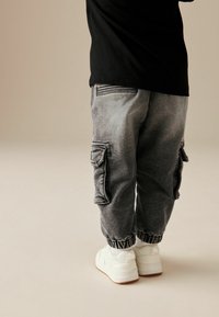 Pantalones cargo grises con puños elásticos, con bolsillos laterales y costuras en contraste, combinados con zapatillas blancas sobre un fondo claro.