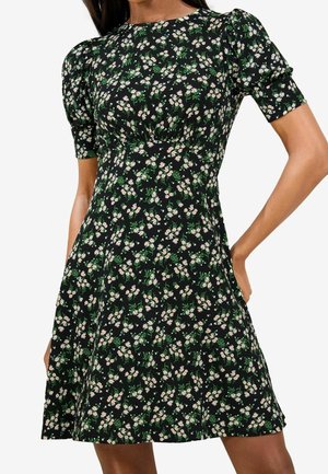 Robe noire à manches courtes avec petit imprimé floral blanc et rose et détail froncé à la taille, portée par une personne debout.