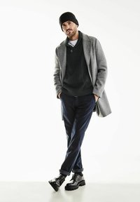 Abrigo de lana gris sobre un suéter de punto oscuro, combinado con pantalones azul marino y botas negras con cordones. El modelo lleva un gorro negro.