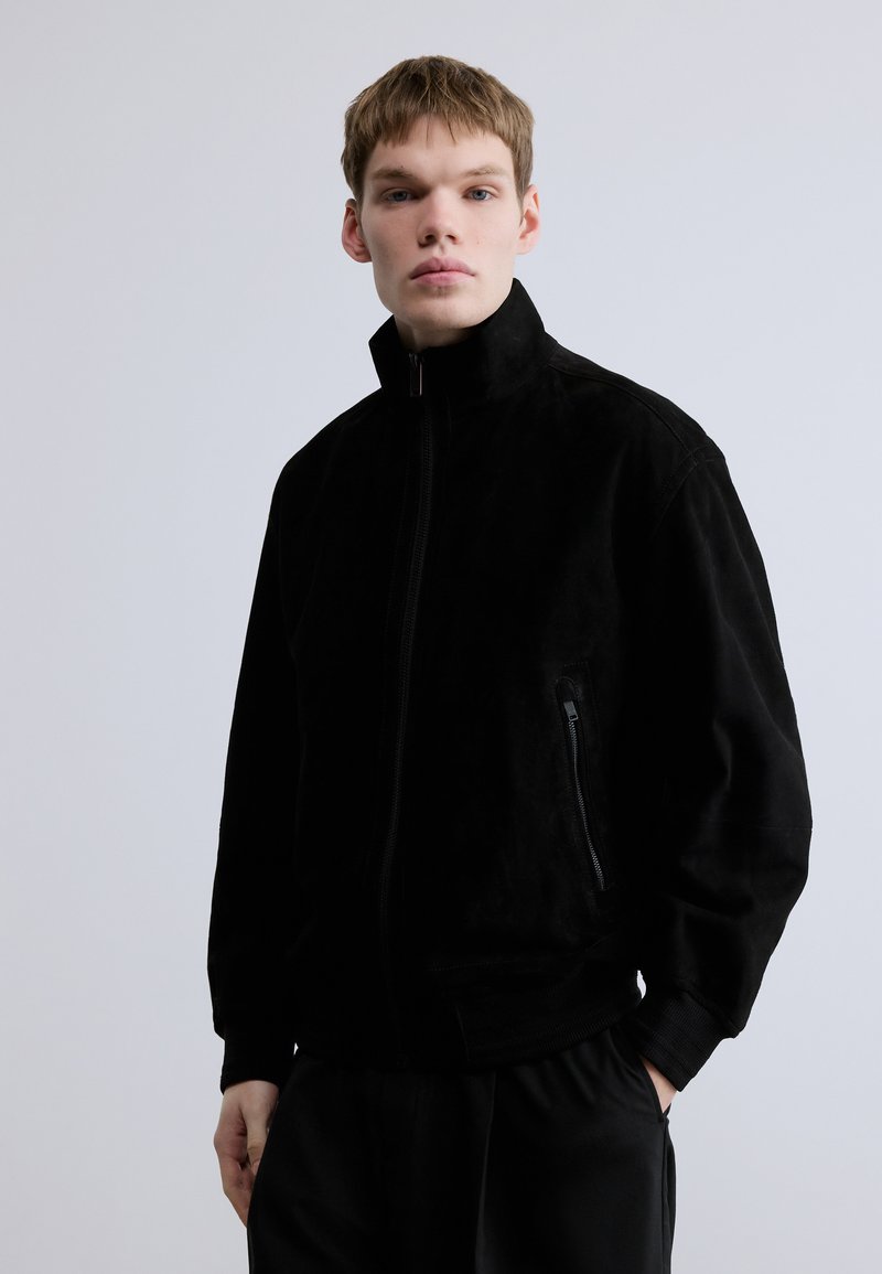 Μαύρο σουέτ μπουφάν με ψηλό γιακά και φερμουάρ μπροστά. Διαθέτει πλαϊνή τσέπη με φερμουάρ και ριμπ μανσέτες. Λείο υλικό με μίνιμαλ σχεδιασμό.