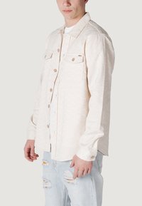 Camicia bianca a maniche lunghe con motivo, dotata di due tasche sul petto e bottoni marroni, indossata sopra una t-shirt bianca e abbinata a jeans azzurri chiari strappati.