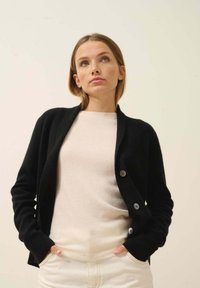 Czarny kardigan z kołnierzem szalowym, miękka tekstura i zapięcie na trzy guziki. Połączony z jasnobeżowym swetrem i kremowymi spodniami.