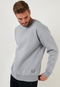 Sudadera gris claro con cuello redondo, puños y dobladillo acanalados. Presenta un pequeño parche de logo negro en la parte inferior izquierda. Textura suave.