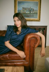 Moteris mėlyname suknelėje, atsilošianti ant rudos odinės sofos, viena ranka remiasi į porankį, fone matyti rėminis paveikslas ir knygų spinta.