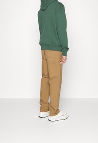 Person klädd i grön huvtröja, beige cargobyxor med fickor i lapp och vita sneakers som står mot en enkel vit bakgrund.