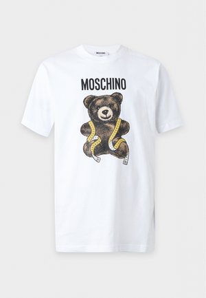 Mladý muž nosí biele tričko Moschino s grafikou medveďa, čierne nohavice so zlatými zipsami a čierne mokasíny, stojí s rukami v vreckách.