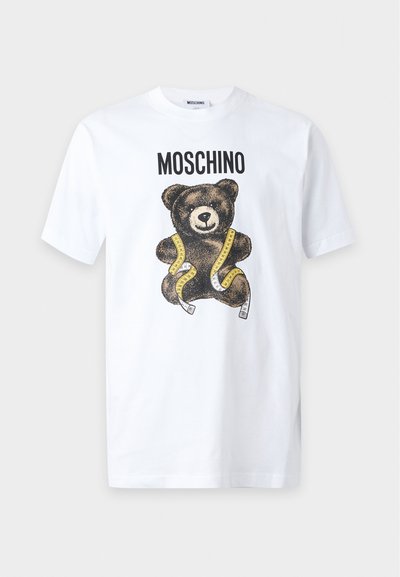 Λευκό κοντομάνικο μπλουζάκι με το κείμενο "MOSCHINO" πάνω από ένα καφέ αρκουδάκι τυλιγμένο με κίτρινη μεζούρα.