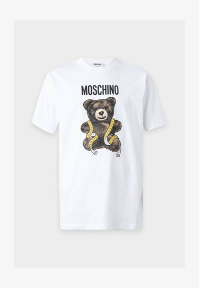 Λευκό κοντομάνικο μπλουζάκι με το κείμενο "MOSCHINO" πάνω από ένα καφέ αρκουδάκι τυλιγμένο με κίτρινη μεζούρα.