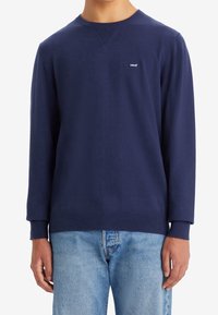 Marinblå sweatshirt gjord av mjukt material, med rund hals, ribbade ärmslut och en liten logotyp på bröstet. Matcher med ljusa jeans.