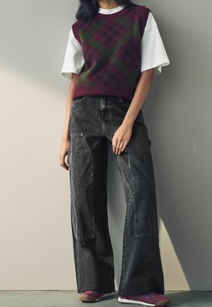 Person trägt ein weißes kurzärmeliges Hemd unter einer lila und grünen gemusterten Strickweste, schwarze Jeans mit weitem Bein und lila Sneakers.