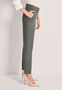 Vrouw in grijze op maat gemaakte broek met zijzakken en beige hoge hakken, staande in profiel tegen een effen achtergrond.