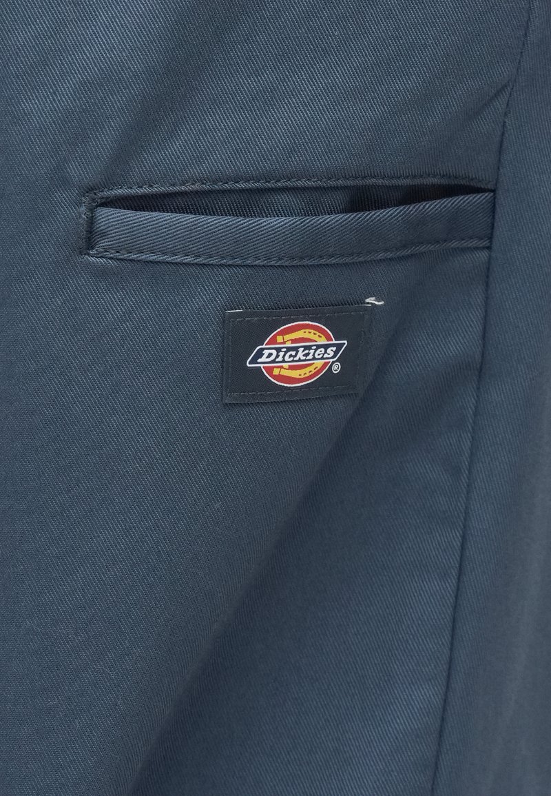 Los pantalones Dickies tienen una tela de color azul oscuro con una textura suave. Un pequeño parche con el logotipo rectangular está colocado en el lateral, cerca de un bolsillo.