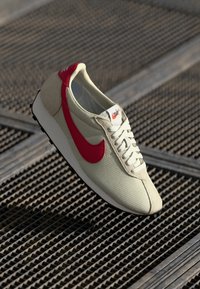 Light beige sneaker with a red Nike swoosh, textured mesh upper, white laces, and contrasting sole. The design features a retro athletic style.