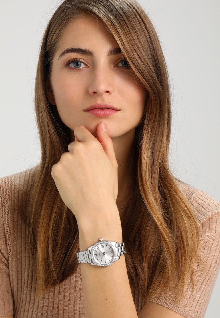 Jeune femme aux longs cheveux châtain clair porte un haut côtelé beige et une montre-bracelet en métal argenté, reposant son menton sur sa main sur un fond uni.