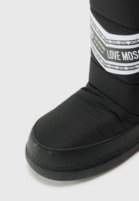 Fekete téli csizma lekerekített orral, bélelt anyagból, és egy fehér csíkkal, amelyen a "LOVE MOSCHINO" márkaneve szerepel. Simább textúra és kontrasztos talp.