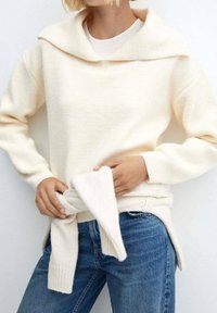 Pull surdimensionné couleur crème avec un col large, des poignets côtelés et un détail à nouer à la taille, associé à un jean en denim bleu.