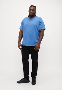 Kék pamut póló rövid ujjakkal, kerek nyakkal és kis piros logóval. Fekete, slim-fit nadrággal és fekete sportcipővel párosítva.
